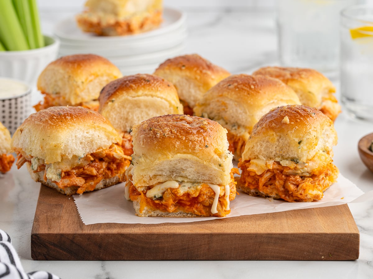 A batch of Buffalo Chicken Sliders filled with shredded chicken and melted cheese sits on a wooden board lined with parchment paper. More sliders and plates are visible in the background, making this Buffalo Chicken Sliders Recipe perfect for sharing.