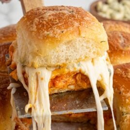 A close-up of a cheesy, golden-brown pull-apart bread roll being lifted, revealing gooey melted cheese and a savory Buffalo Chicken Sliders filling inside. Other similar rolls are visible in the background.