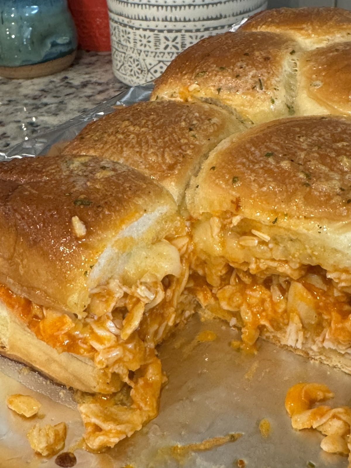 A close-up of Buffalo Chicken Sliders filled with shredded chicken in an orange sauce, likely made from a classic Buffalo Chicken Sliders Recipe. One slider is removed, revealing a cheesy filling and the golden-brown, seasoned top.