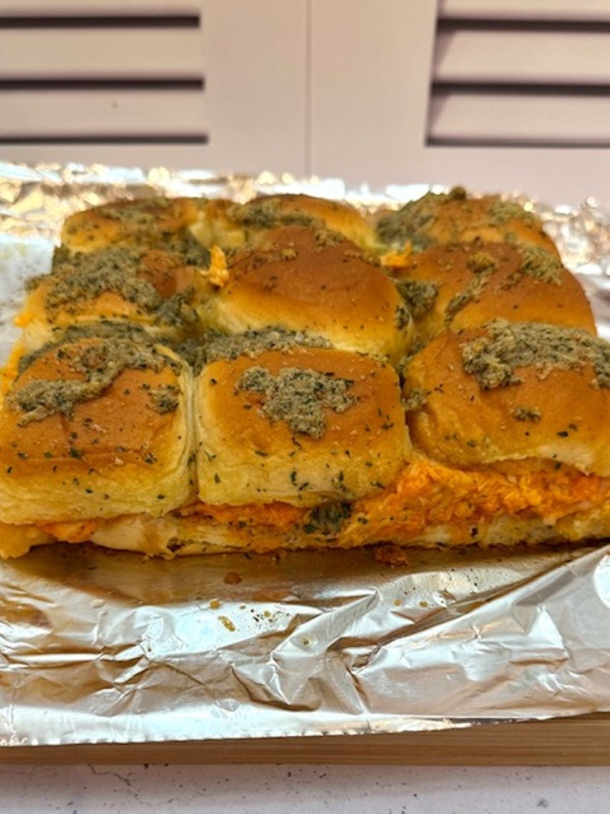 A tray of Buffalo Chicken Sliders with golden brown tops, seasoned with herbs and butter, filled with a creamy orange filling, sits on aluminum foil.