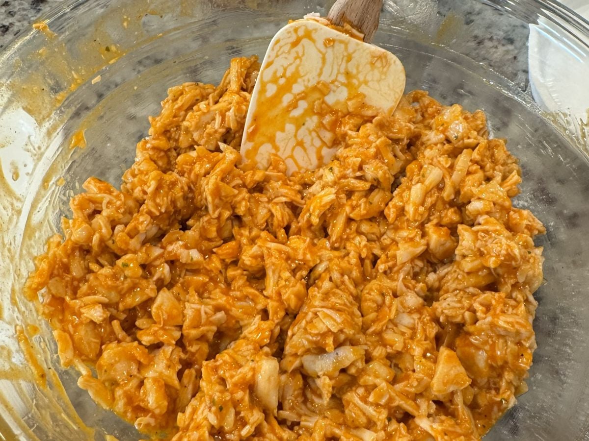 A glass bowl filled with shredded chicken mixed with a tangy orange sauce, being stirred with a white spatula&mdash;perfect for your next Buffalo Chicken Sliders Recipe.