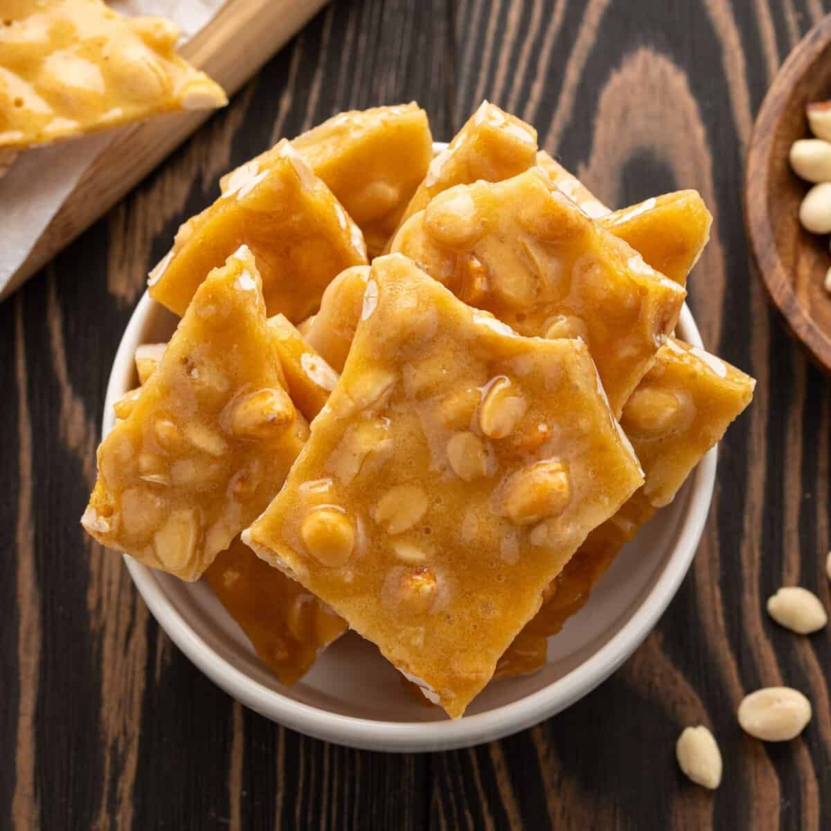 A bowl filled with pieces of Microwave Peanut Brittle sits on a wooden table, with some peanuts and more brittle visible in the background.