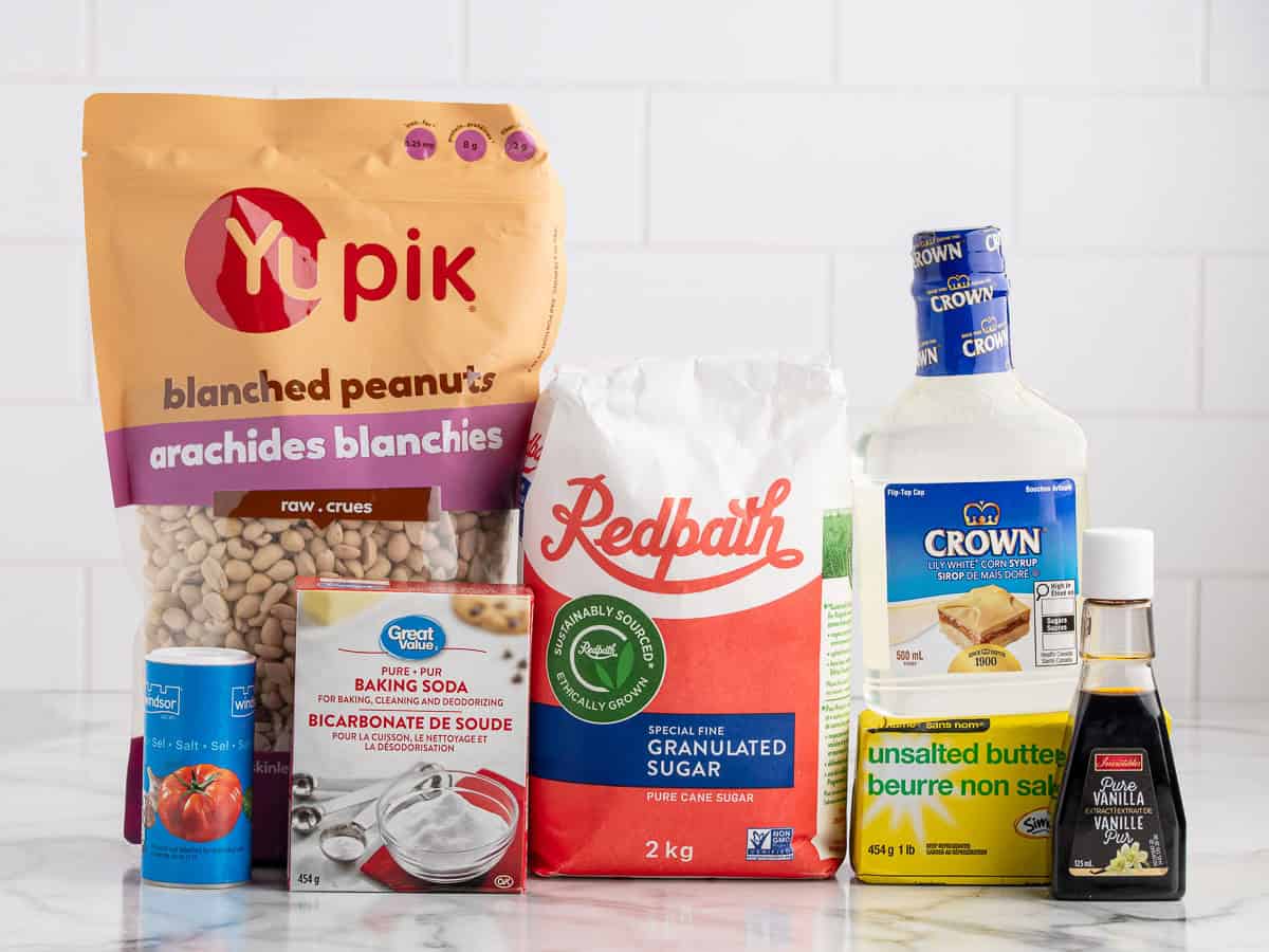 Assorted baking ingredients on a counter, perfect for making Microwave Peanut Brittle, including blanched peanuts, sugar, corn syrup, baking soda, butter, vanilla extract, and a box of baking soda, in front of a white tiled wall.