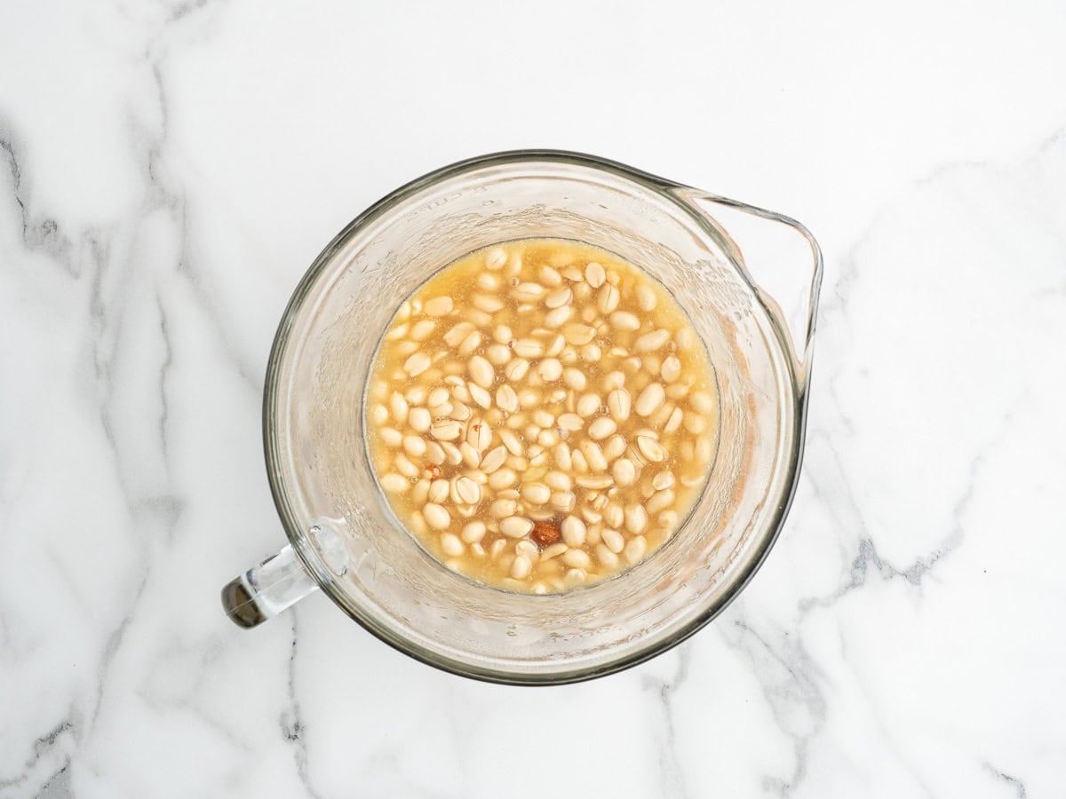 A glass measuring cup filled with white beans soaking in water on a white marble surface, reminiscent of preparing ingredients for recipes like Microwave Peanut Brittle.