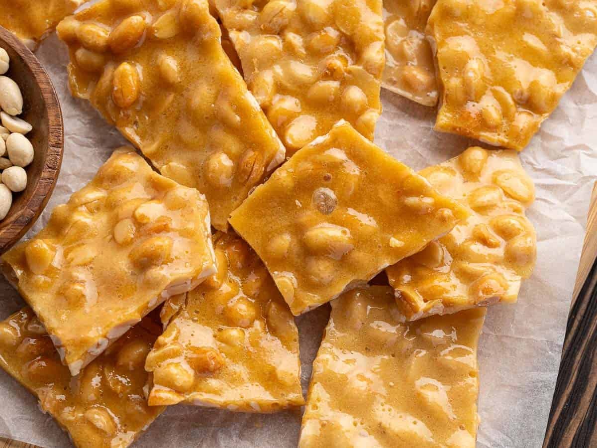 Close-up of pieces of Microwave Peanut Brittle on parchment paper, showing glossy, golden-brown candy with whole peanuts inside. A bowl of peanuts is partially visible in the upper left corner of the image.