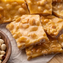 Close-up of Microwave Peanut Brittle pieces on parchment paper, with a wooden bowl of shelled peanuts to the side. The brittle is golden brown and glossy, showing embedded peanuts.