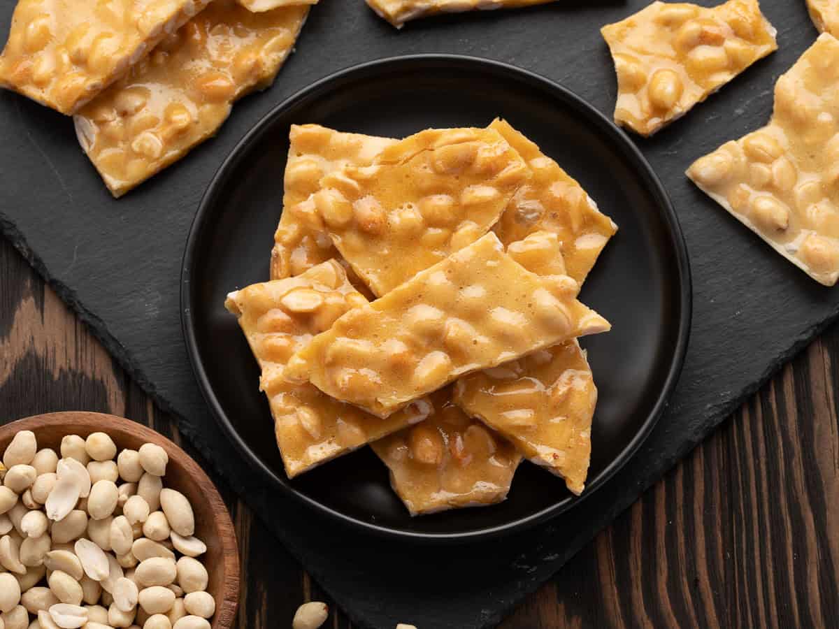 A black plate with pieces of Microwave Peanut Brittle sits on a dark wooden table. A bowl of shelled peanuts and more brittle pieces are nearby on a slate board.