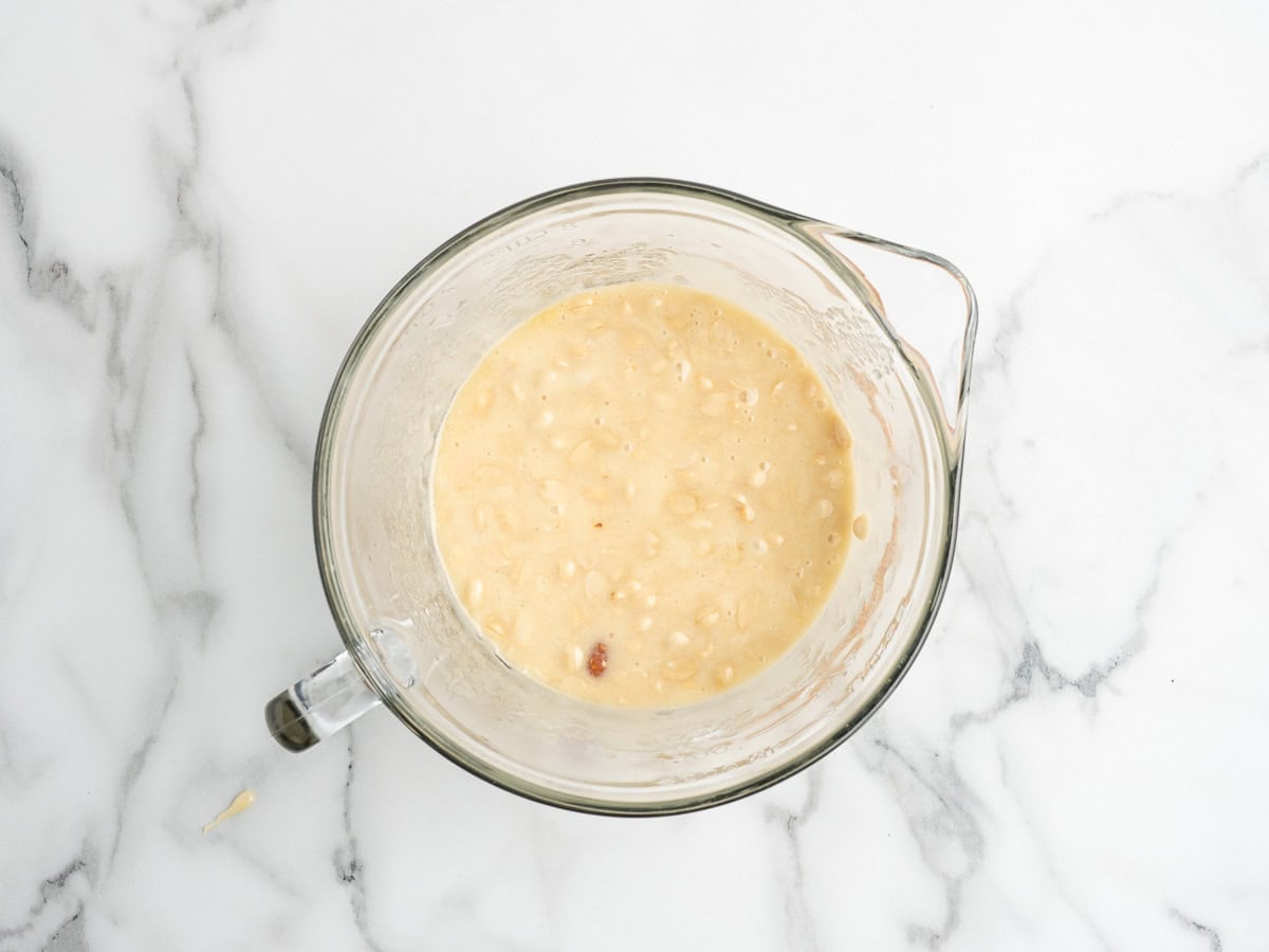 A glass mixing bowl filled with lumpy banana bread batter sits on a white marble countertop, next to a plate of homemade Microwave Peanut Brittle.