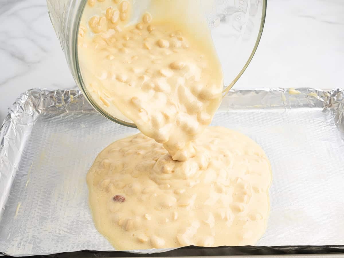 A thick, nut-filled batter for Microwave Peanut Brittle is being poured from a glass bowl onto a foil-lined baking sheet.