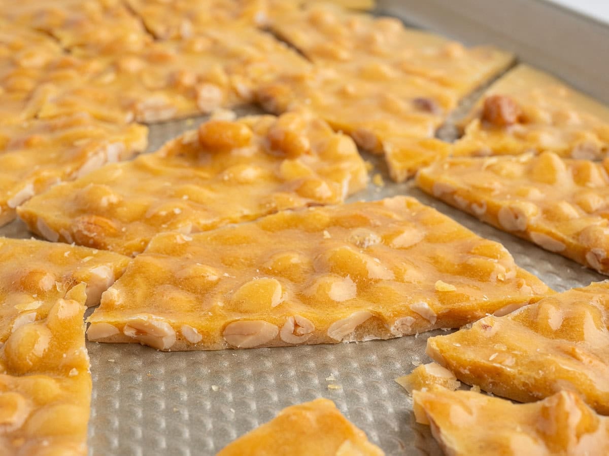 Close-up of pieces of Microwave Peanut Brittle on a textured metal baking sheet, showing golden caramelized sugar and embedded peanuts.