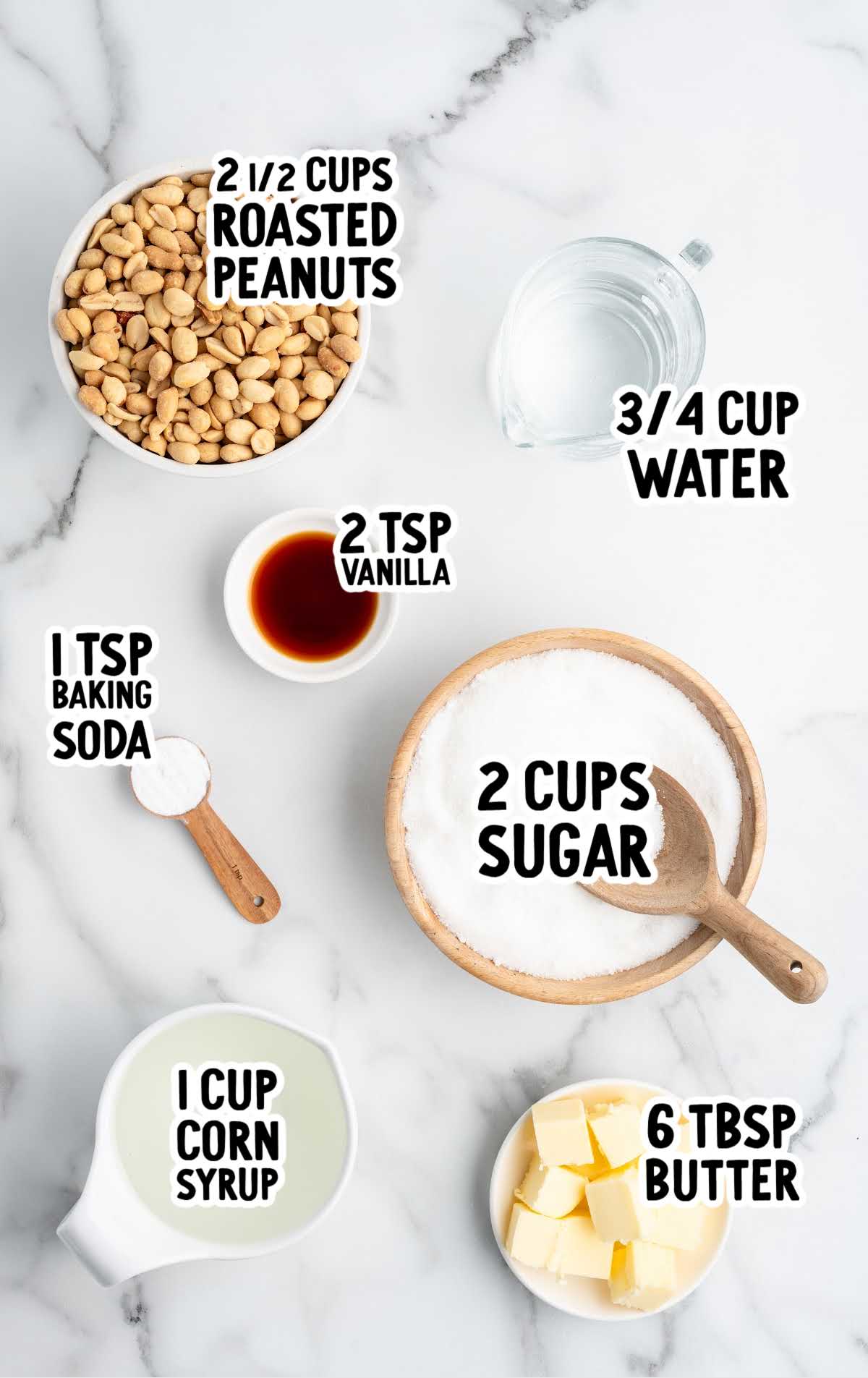 Peanut brittle ingredients on a marble counter including sugar, corn syrup, butter, roasted peanuts, vanilla, baking soda, and water