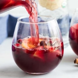 Fall sangria being poured from a pitcher into a stemless wine glass, surrounded by fruit and cinnamon sticks