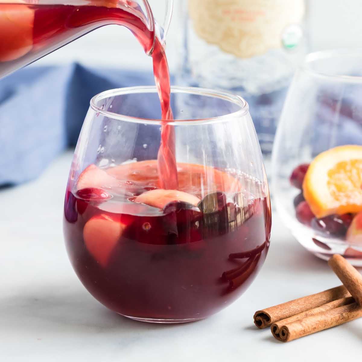 Square image of sangria being poured into a glass filled with apples, cranberries, and cinnamon sticks
