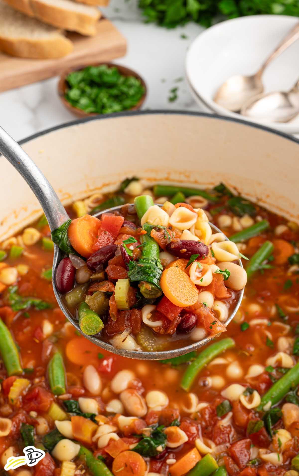 Ladle full of vibrant minestrone soup above a pot, showing detailed textures of beans, vegetables, and small shell pasta.
