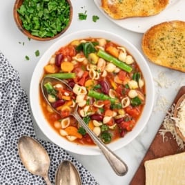 Recipe card style photo of minestrone soup in a white bowl with vintage spoons, fresh parsley, and toasted bread arranged around it.