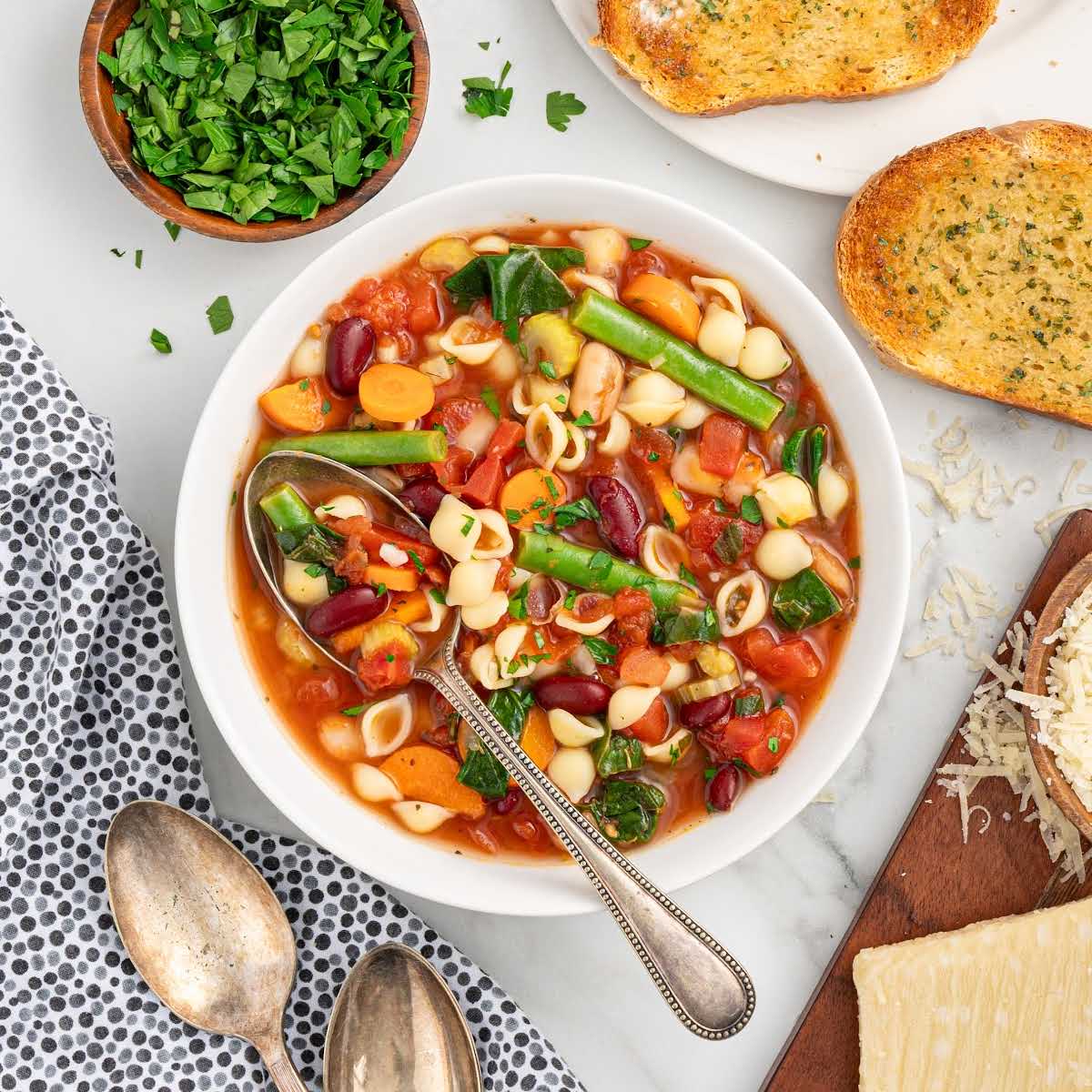 Recipe card style photo of minestrone soup in a white bowl with vintage spoons, fresh parsley, and toasted bread arranged around it.