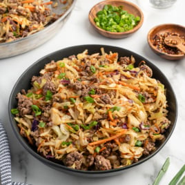 A black bowl filled with a Mongolian Beef stir-fry&mdash;ground beef, shredded cabbage, carrots, and green onions&mdash;sits on a marble surface. Nearby are small bowls of green onions, chili flakes, and a pan with more Mongolian Beef Recipe.