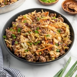 A black bowl filled with a Mongolian Beef stir-fry&mdash;ground beef, shredded cabbage, carrots, and green onions&mdash;sits on a white surface, with small bowls of sliced green onions and chili flakes nearby for this easy Mongolian Beef recipe.