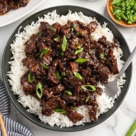 A bowl of white rice topped with glazed Mongolian Beef strips, garnished with sliced green onions. A fork rests on the bowl, and a small dish of extra green onions is nearby.