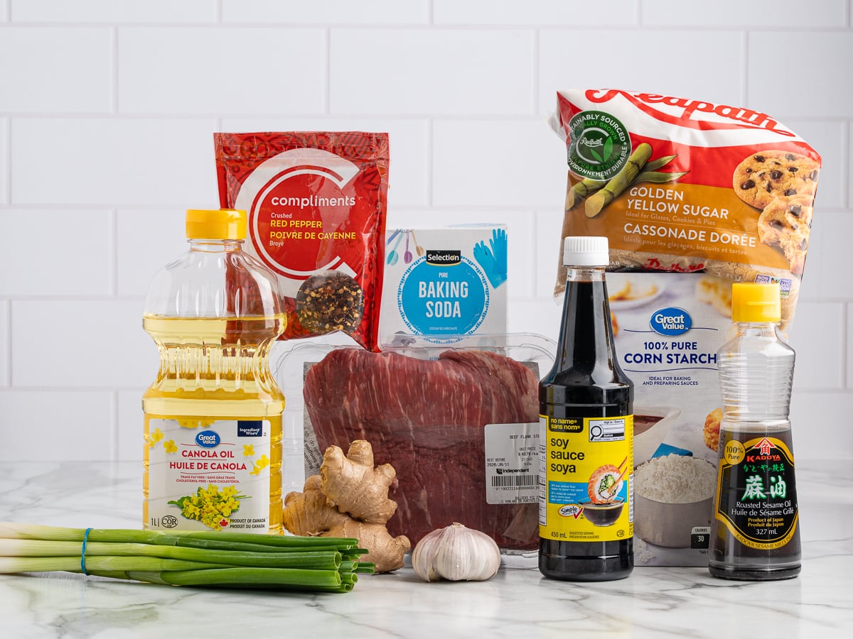 A variety of cooking ingredients on a marble counter, perfect for a Mongolian Beef Recipe, including canola oil, ginger, garlic, green onion, soy sauce, sesame oil, baking soda, cornstarch, brown sugar, red pepper flakes, beef, and white rice.