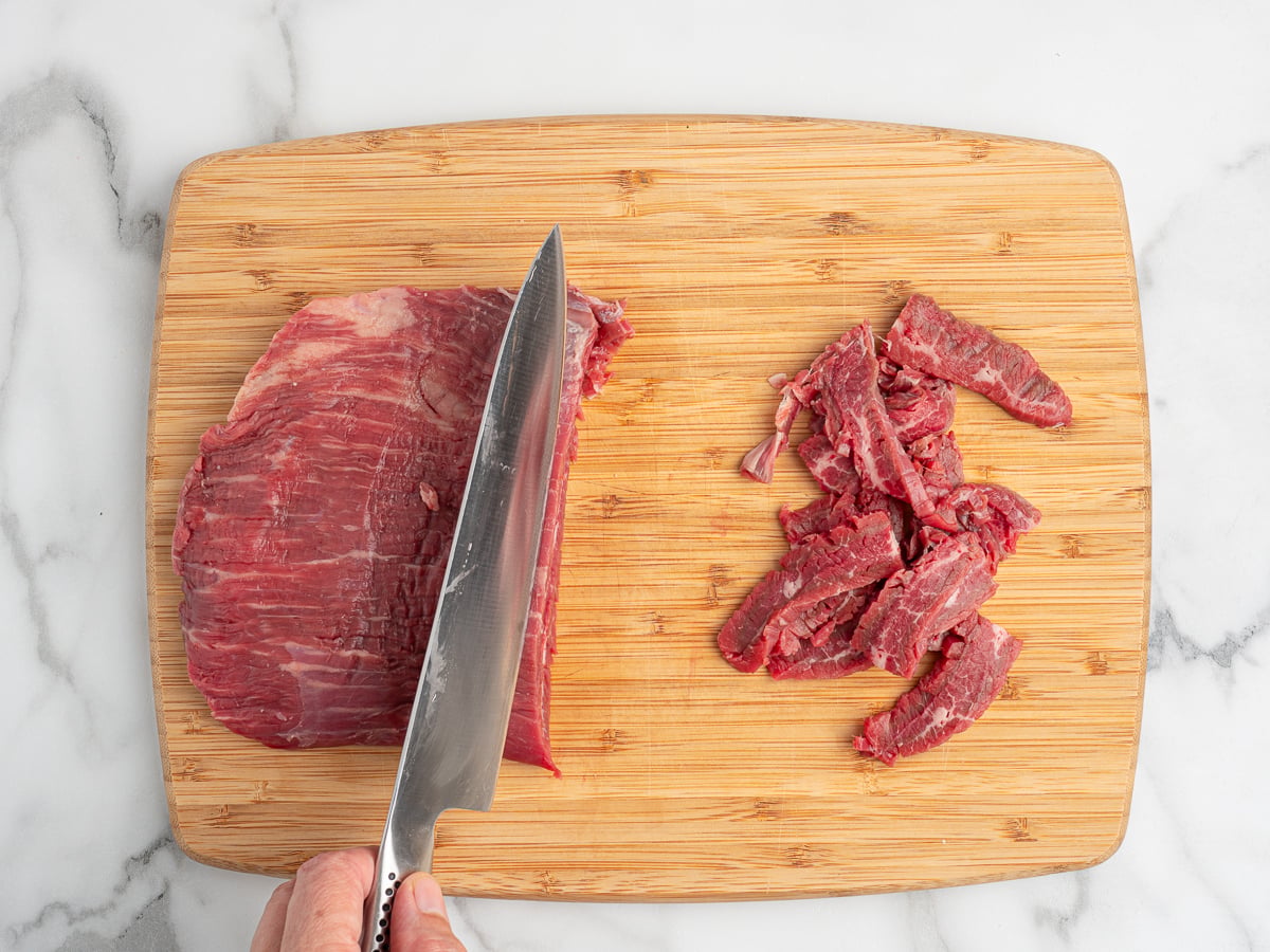 A hand uses a large knife to slice raw beef, perfect for a Mongolian Beef recipe, on a wooden cutting board. Thin strips are arranged beside the uncut portion on a marble countertop.