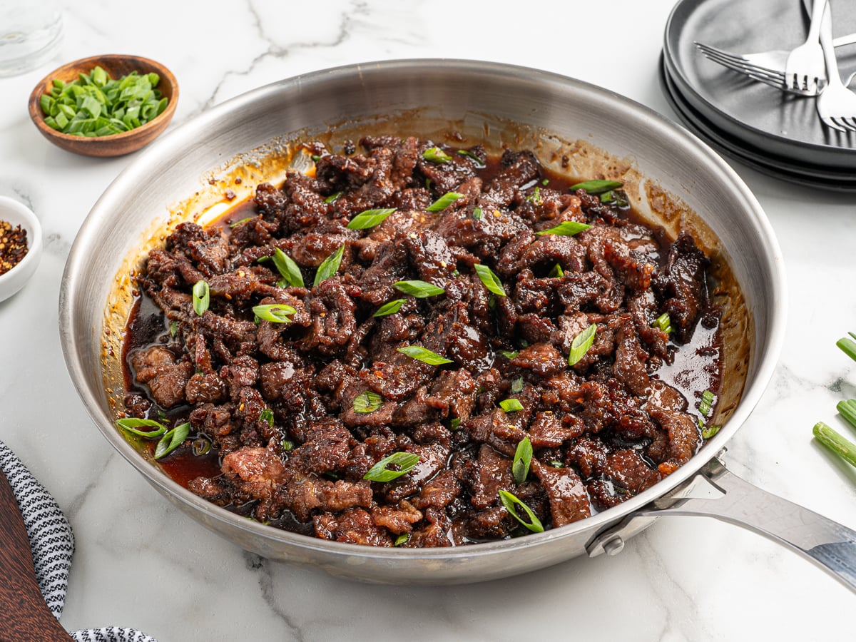 A stainless steel skillet filled with saucy Mongolian Beef, topped with sliced green onions, sits on a marble surface. Plates, utensils, and a bowl of chopped green onions are nearby&mdash;perfect for serving this delicious Mongolian Beef Recipe.