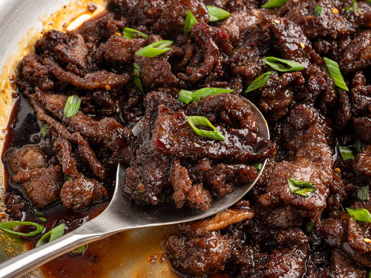 A close-up of caramelized beef strips in a rich, dark Mongolian Beef sauce, garnished with sliced green onions, with a metal spoon scooping some of the beef.