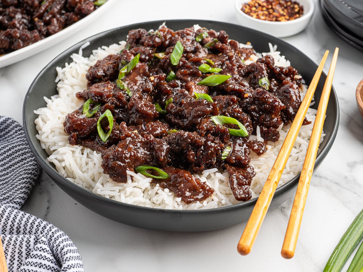 A black bowl filled with white rice topped with glazed Mongolian Beef and garnished with sliced green onions, with chopsticks resting on the side. A striped napkin and small bowl of red pepper flakes complement this delicious Mongolian Beef Recipe.