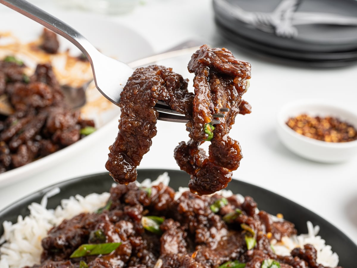 A close-up of a fork holding saucy Mongolian Beef strips above a plate of white rice, garnished with green onions. In the background, more Mongolian Beef, rice, and a small bowl of red chili flakes are visible.