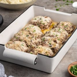 A white baking dish holds eight cooked Salisbury steaks—often called Poor Man’s Steak—topped with creamy mushroom gravy and chopped parsley. The dish sits on a gray countertop with a bowl of mashed potatoes and a small bowl of herbs nearby.