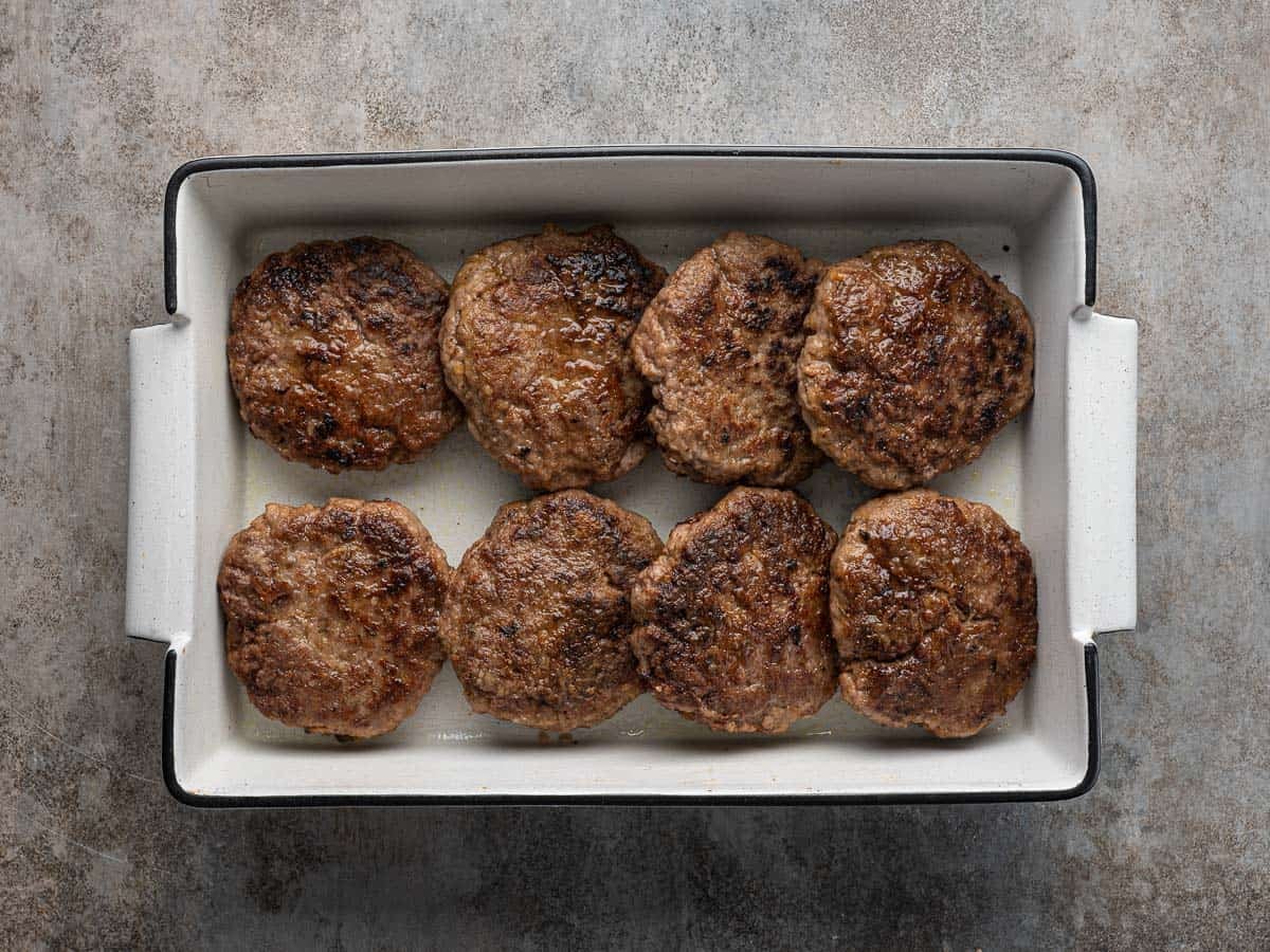 Eight browned beef patties, reminiscent of a classic Poor Man’s Steak, are arranged in two neat rows inside a white rectangular baking dish, placed on a textured gray surface.