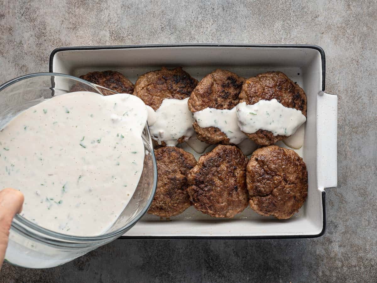 A hand pours creamy white sauce with herbs over browned meat patties arranged in a white rectangular baking dish on a gray surface, creating a classic Poor Man’s Steak Recipe that’s both comforting and delicious.