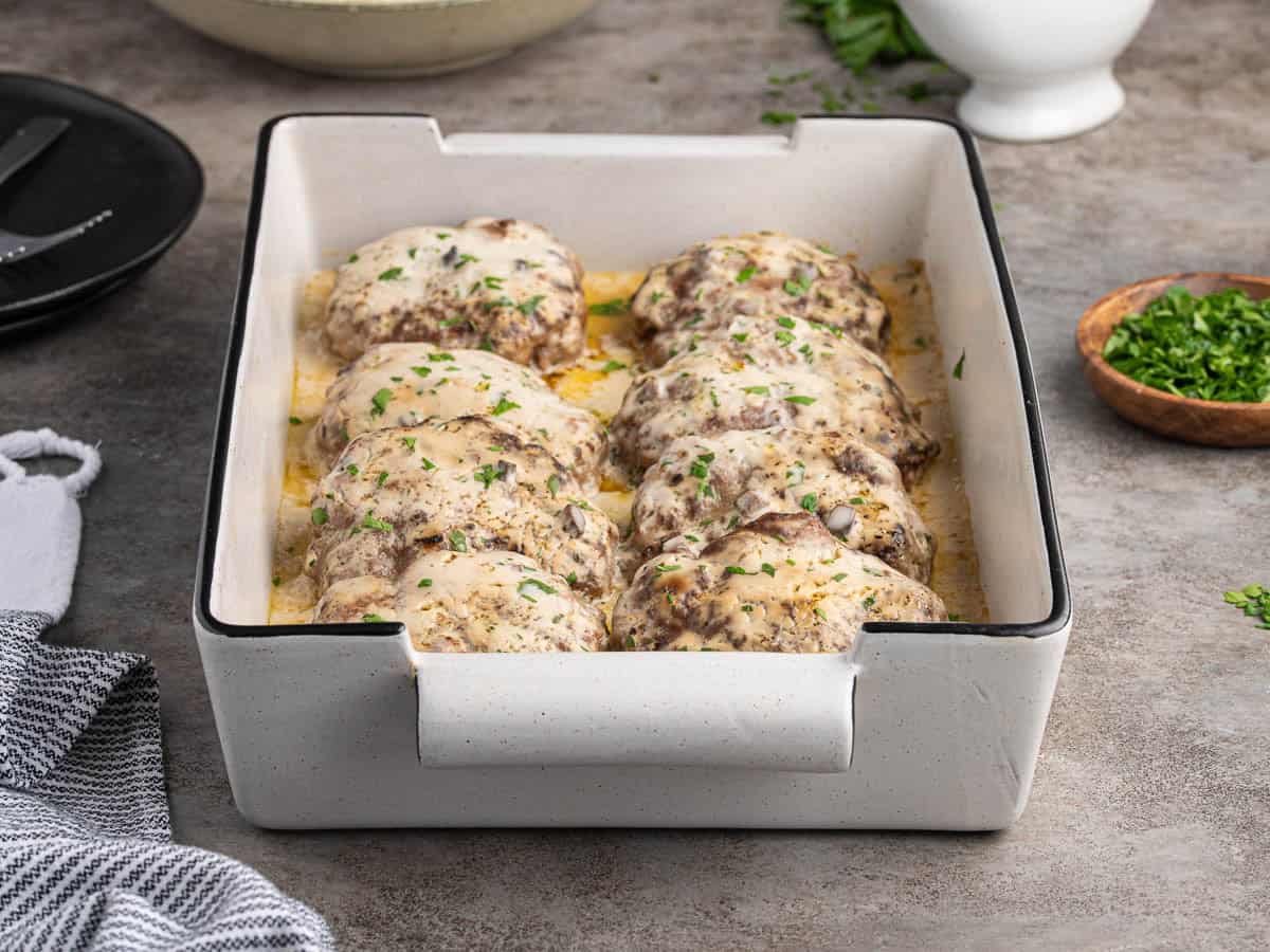 A white baking dish contains eight baked chicken thighs topped with melted cheese and herbs, garnished with chopped parsley. Reminiscent of a classic Poor Man’s Steak Recipe, this hearty dish sits on a gray countertop near a towel and a bowl of extra parsley.