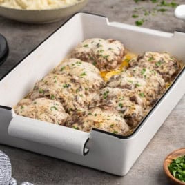 A white baking dish with eight baked meat patties, inspired by a classic Poor Man’s Steak Recipe, topped with melted cheese and chopped herbs, sitting on a gray countertop next to a striped towel and a bowl of fresh parsley.