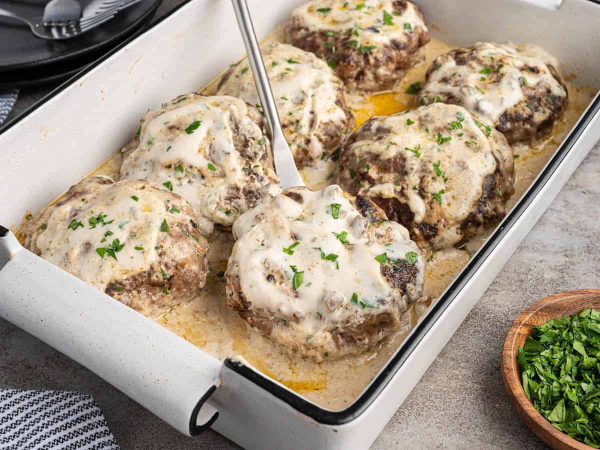 Six baked Poor Man’s Steaks in a white pan, topped with creamy sauce and chopped parsley. A metal spatula is lifting one steak. A small bowl of more parsley is on the side, complementing this classic Poor Man’s Steak recipe.