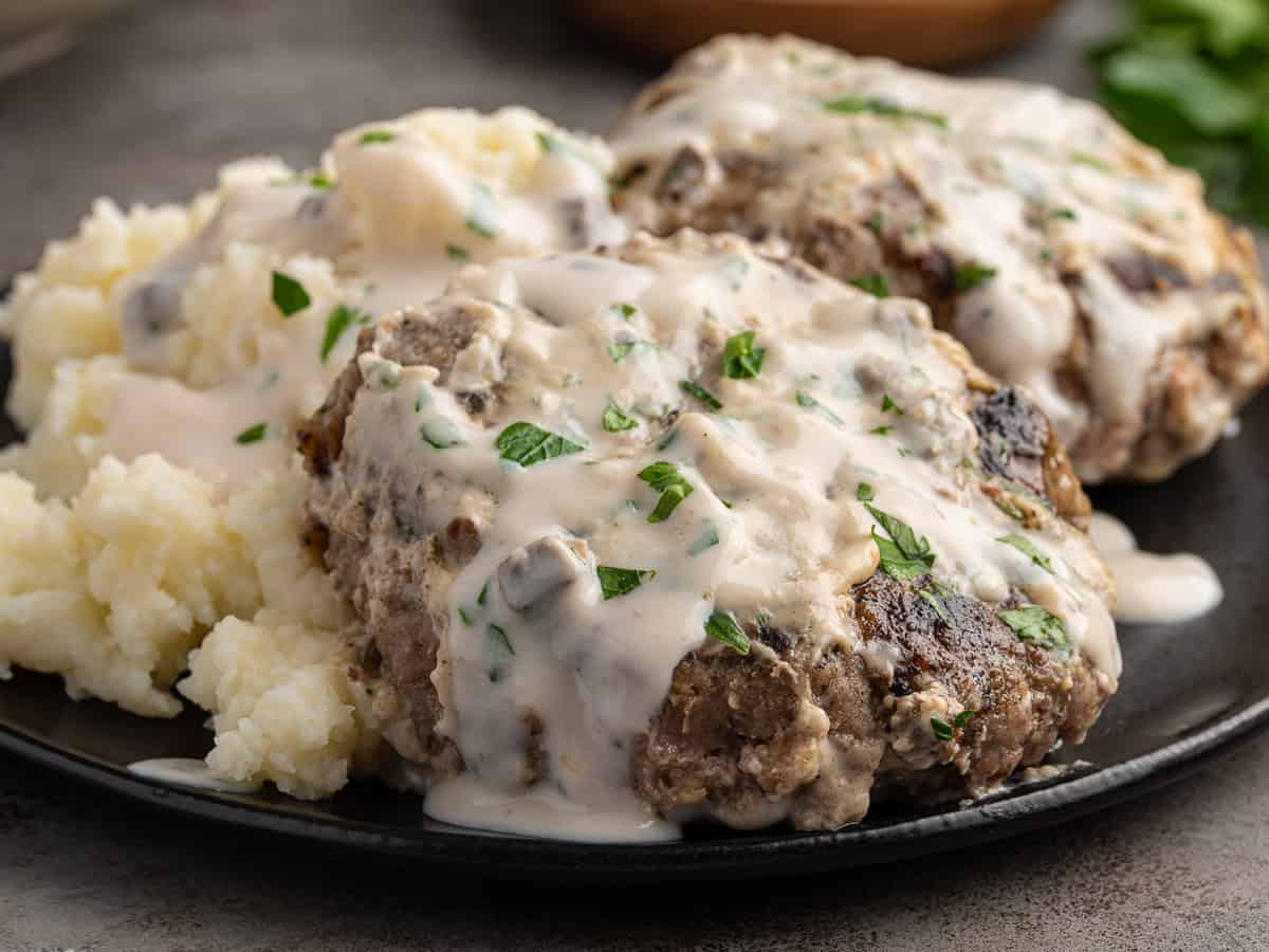 Two hamburger steaks, inspired by a classic Poor Man’s Steak Recipe, are covered in creamy mushroom gravy and garnished with chopped parsley, served next to a helping of mashed potatoes on a dark plate.