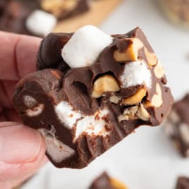 A hand holds a piece of Rocky Road Fudge, showcasing chocolate, mini marshmallows, and peanuts.