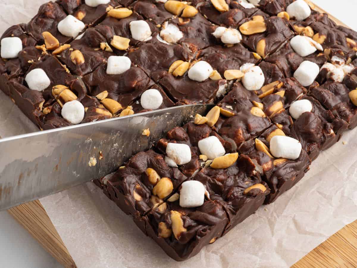 A knife slices through a block of Rocky Road Fudge topped with peanuts and mini marshmallows, placed on parchment paper over a wooden cutting board.