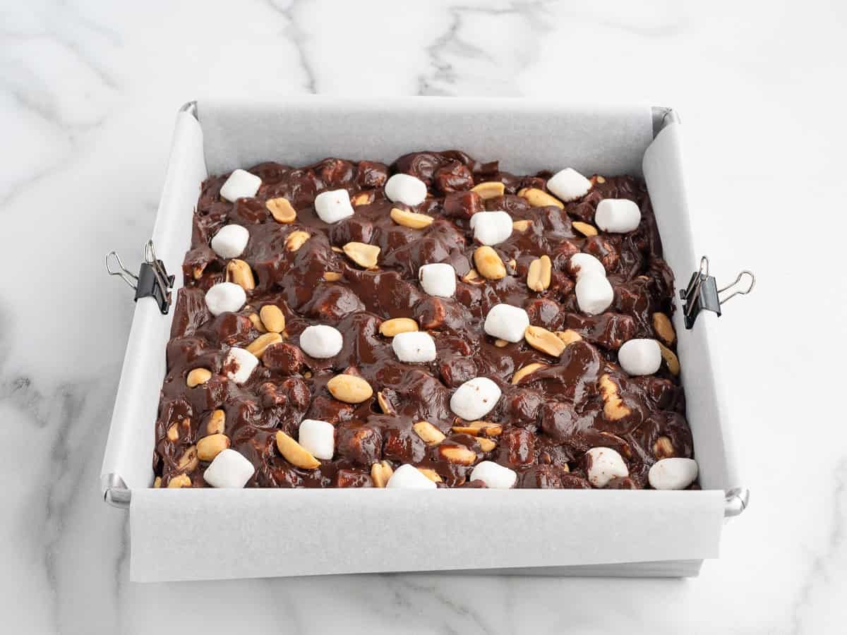 A square baking pan lined with parchment paper holds delicious Rocky Road Fudge topped with marshmallows and peanuts, sitting on a white marble surface.
