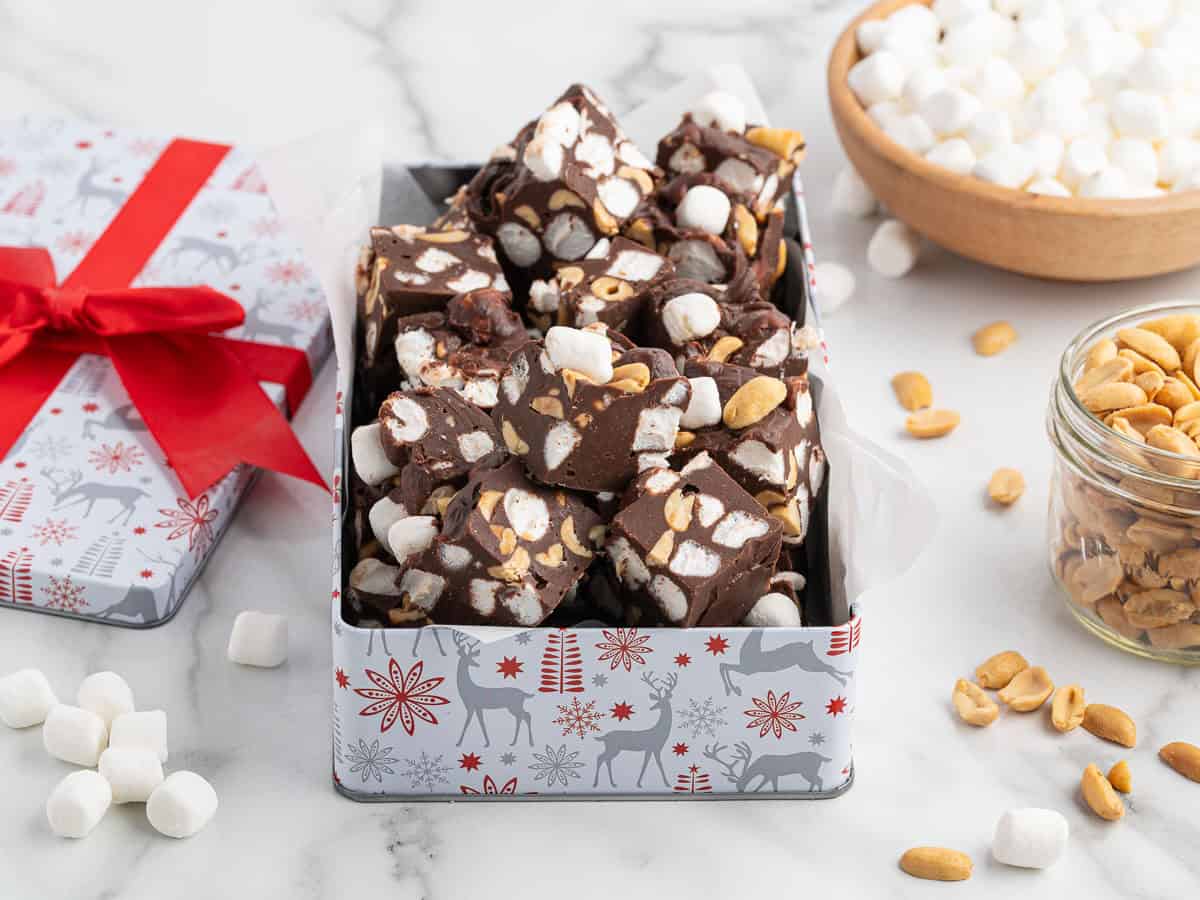 A decorative tin filled with Rocky Road Fudge—chocolate fudge pieces containing marshmallows and peanuts—is surrounded by a bowl of marshmallows, a jar of peanuts, and scattered marshmallows on a marble surface.