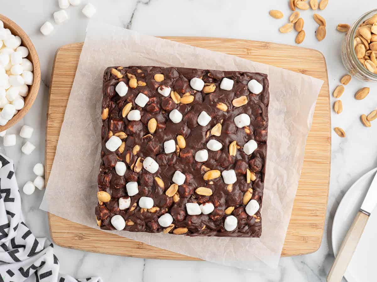 A square chocolate dessert, made from a classic Rocky Road Fudge recipe, is topped with peanuts and mini marshmallows as it sits on parchment paper on a wooden board, surrounded by bowls of peanuts and marshmallows.
