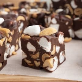 Squares of Rocky Road Fudge filled with mini marshmallows and peanuts are arranged on parchment paper. The treats have a rich, creamy texture, and the nuts and marshmallows are visibly mixed throughout.