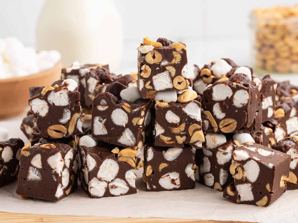 A stack of decadent Rocky Road Fudge pieces filled with peanuts and mini marshmallows sits on parchment paper, with bowls of marshmallows and peanuts blurred in the background—perfect for anyone looking to try a classic Rocky Road Fudge Recipe.