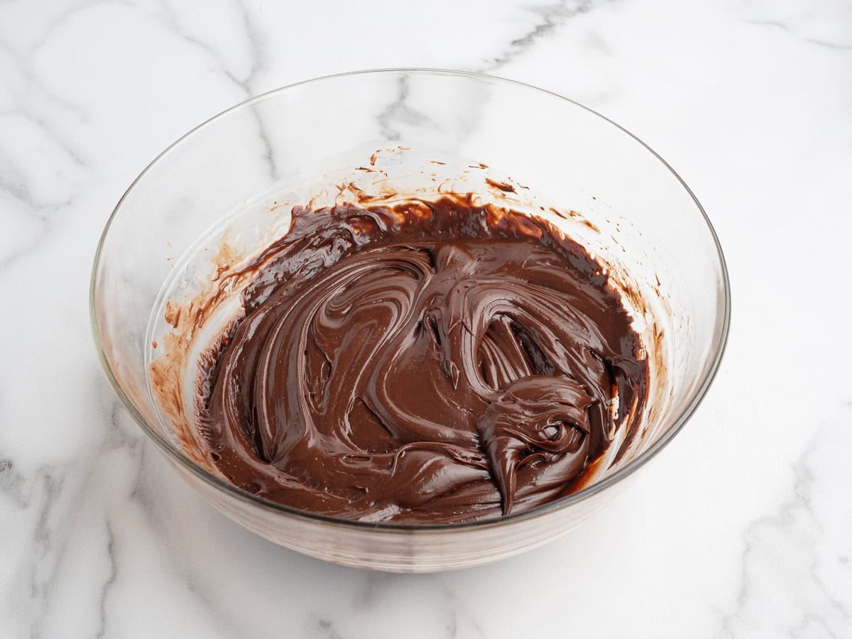 A clear glass bowl filled with smooth, swirled chocolate batter sits on a white marble surface, ready to be transformed into a decadent Rocky Road Fudge Recipe.
