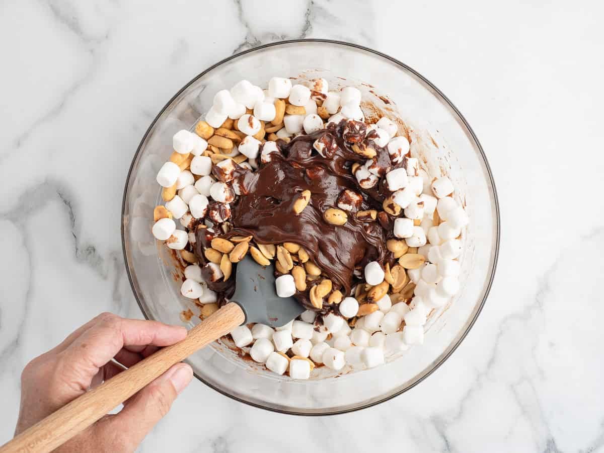 A hand stirs mini marshmallows, peanuts, and melted chocolate in a glass bowl with a spatula on a marble countertop, preparing a classic Rocky Road Fudge.
