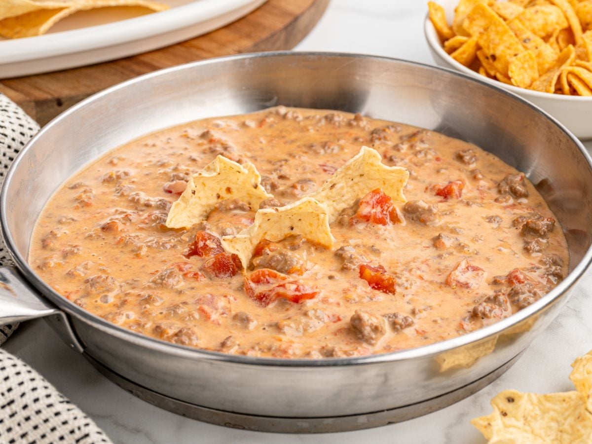 A metal skillet filled with creamy ground beef and tomato Rotel Dip, garnished with three tortilla chips standing upright. A bowl of corn chips and a plate of tortillas are visible in the background.