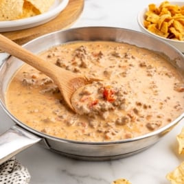 A skillet filled with creamy Rotel Dip, featuring ground beef and tomatoes, is being stirred with a wooden spoon. Tortilla chips and corn chips are arranged around the skillet on a white surface.