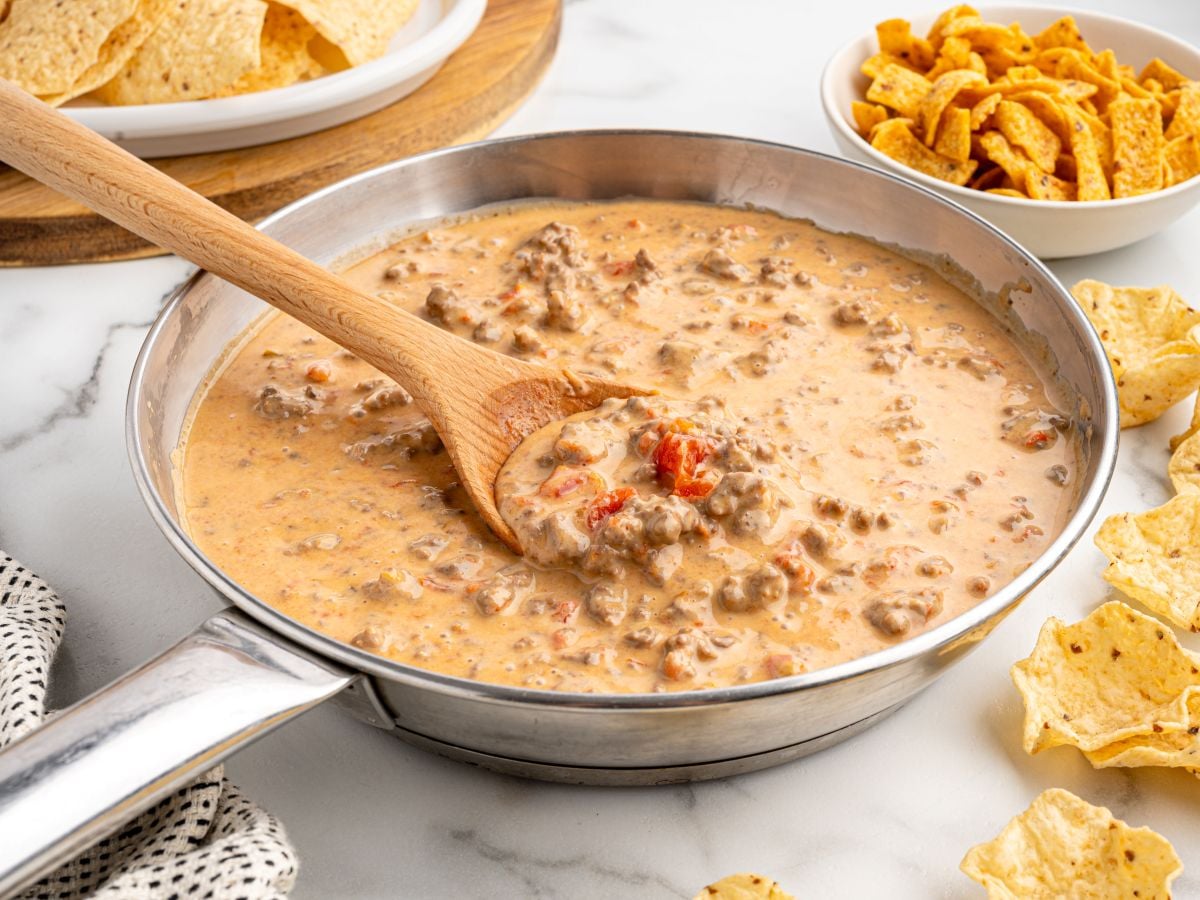 A skillet filled with creamy Rotel Dip, featuring ground beef and tomatoes, is being stirred with a wooden spoon. Tortilla chips and corn chips are arranged around the skillet on a white surface.