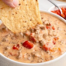 A hand dips a large tortilla chip into a bowl of creamy Rotel Dip with ground beef and diced tomatoes. In the background, a small bowl of salsa and more chips are visible.