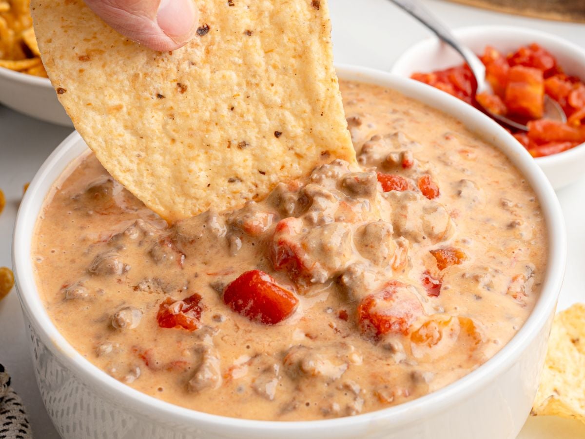 A hand dips a large tortilla chip into a bowl of creamy Rotel Dip with ground beef and diced tomatoes. In the background, a small bowl of salsa and more chips are visible.